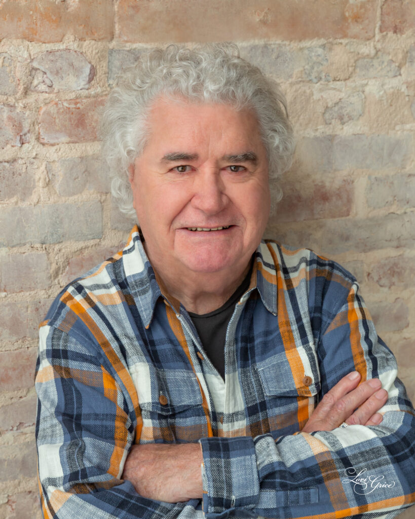 A man with white hair and plaid shirt standing in front of brick wall.